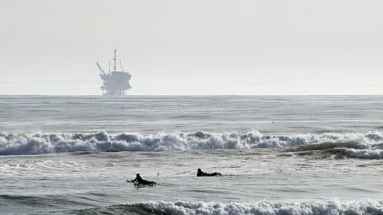 Two surfers paddle past the breaking waves, an oil outrigger looms in the distance.